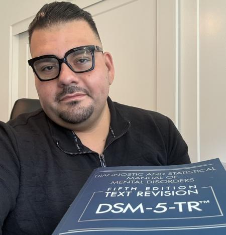 Professor Jose Luna holding a DSM-5-TR (Diagnostic and Statistical Manual of Mental Disorders) textbook.